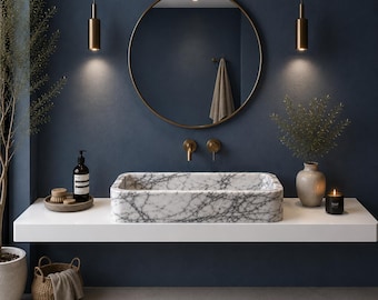 May include: A rectangular white marble sink with dark grey veining sits on a floating white shelf. Above the sink is a round mirror with a thin gold frame, flanked by two gold pendant lights. A gold faucet is mounted on the dark blue wall.