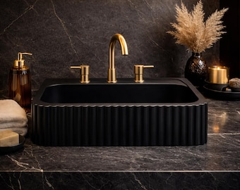 Könnte beinhalten: Ein rechteckiges schwarzes Waschbecken mit vertikal geriffeltem Außendesign. Das Waschbecken verfügt über eine hohe, geschwungene goldene Armatur und zwei passende goldene Griffe auf beiden Seiten. Das Becken ist tief, mattschwarz und metallisch.