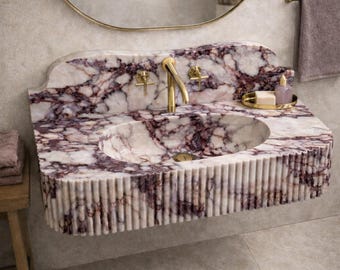 Könnte beinhalten: Ein wandmontiertes Waschbecken aus weißem, dunkelviolett geädertem Marmor. Das Becken verfügt über ein geriffeltes Außendesign mit vertikalen Rillen und ist mit einer goldfarbenen Armatur sowie zwei passenden Kreuzgriffventilen ausgestattet.