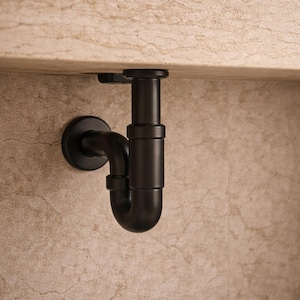 May include: A matte black P-trap plumbing fixture featuring a curved pipe design with a circular wall flange and a vertical connection point. The metal pipe assembly is finished in a solid dark tone and is mounted to a textured beige surface.