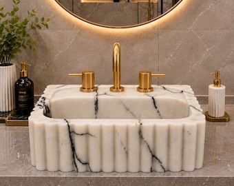 Könnte beinhalten: Ein rechteckiges Waschbecken aus weißem Marmor mit markanter schwarzer Maserung und geriffeltem Außendesign. Das Waschbecken ist mit einer Armatur in gebürstetem Gold und zwei passenden Hebelgriffen ausgestattet, montiert auf einer grauen Steinplatte.