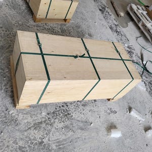 May include: A rectangular wooden shipping crate constructed from light-colored plywood panels. The crate is secured with two dark green plastic strapping bands that wrap around the length and width, fastened with metal buckles on the top surface.