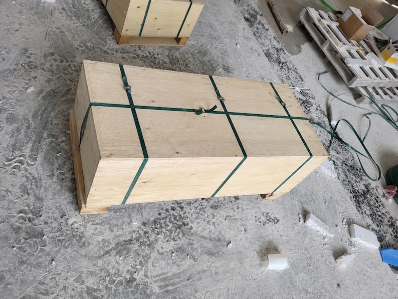 May include: A rectangular wooden shipping crate constructed from light-colored plywood panels. The crate is secured with two dark green plastic strapping bands that wrap around the length and width, fastened with metal buckles on the top surface.