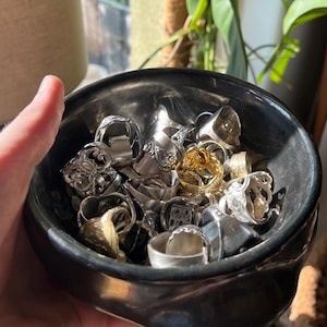 May include: A black ceramic bowl filled with a collection of assorted metal rings. The rings feature various designs, including intricate floral patterns, smooth bands, and textured surfaces, with a mix of silver-toned and gold-toned finishes.