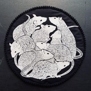 Op de afbeelding: Een zwart, cirkelvormig decoratief item met een witte illustratie van verschillende ratten. De ratten zijn afgebeeld met gedetailleerde vacht en lange staarten. De rand van het item is afgewerkt met een zwarte pomponrand.
