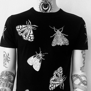 Könnte beinhalten: Schwarzes T-Shirt mit einem weißen Grafikdruck von fünf Motten. Die Motten sind in verschiedenen Größen und Formen und in einem zufälligen Muster auf dem Shirt angeordnet.