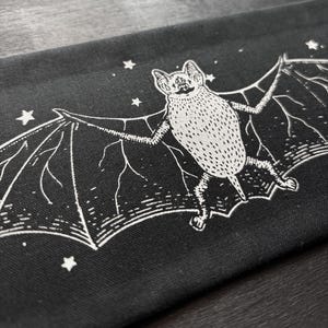 Op de afbeelding: Zwarte stof met een witte illustratie van een vleermuis met uitgespreide vleugels en sterren. De vleermuis is gedetailleerd met getextureerde schaduwen en ingewikkelde vleugelpatronen, wat een opvallend contrast creëert.