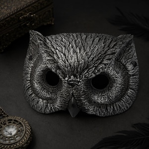 May include: A silver and black owl mask featuring highly detailed, textured feather patterns across the surface. The mask has two large, hollow circular eye openings and a pointed beak, with small holes visible on the sides for attachment.