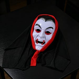 May include: A plastic vampire mask featuring a pale white face, arched black eyebrows, red eye rims, and sharp red fangs. The mask is attached to a black fabric hood with a bright red trim that frames the face, creating a classic horror character look.