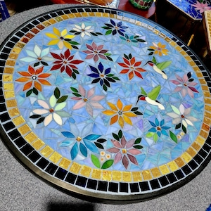 Peut inclure: Un plateau de table rond en mosaïque présentant un fond bleu clair avec diverses fleurs en verre multicolores et deux colibris. La bordure se compose d'un anneau de carreaux dorés entouré d'un anneau extérieur plus large de carreaux noirs.
