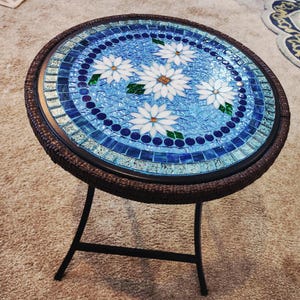 May include: Round mosaic side table with a blue and white floral design. The tabletop features a mosaic of blue tiles with white daisy-like flowers. The table has a dark brown woven edge and a black metal base.