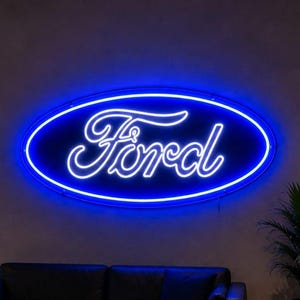 Peut inclure: Une enseigne néon bleu vif affichant le logo Ford en écriture cursive dans un cadre ovale, montée sur un mur gris au-dessus de deux canapés en cuir noir dans un showroom avec des sols polis et plusieurs voitures de sport garées en arrière-plan.