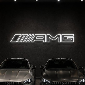 Op de afbeelding: Vier luxe Mercedes-AMG sedans geparkeerd in een showroom met een groot verlicht AMG-logo op de donkere muur. De voertuigen zijn uitgevoerd in zwarte, zilveren en matzwarte afwerkingen, met verticale grille-lamellen en heldere led-koplampen.