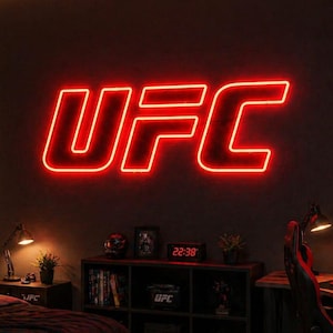 May include: A bedroom featuring a large, glowing red neon UFC sign mounted on a dark wall. The room includes a bed with black bedding, a black shelving unit, a desk with a computer setup, and a poster of a fighter on the wall with the text DISCIPLINE FOCUS RESPECT STRENGTH BELIEF.