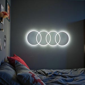 Könnte beinhalten: Ein wandmontiertes Neonschild mit vier ineinandergreifenden weißen kreisförmigen Ringen, die in einer horizontalen Reihe angeordnet sind. Das helle weiße Licht strahlt sanft gegen die dunkle Wand und erzeugt ein geometrisches Muster.