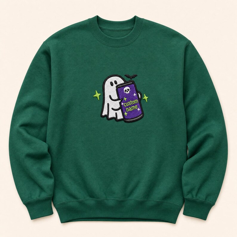 May include: A dark green crewneck sweatshirt featuring an embroidered design of a white ghost holding a purple beverage can. The can displays a skull icon, the text "custom name," and small star accents. A small black bat is positioned above the can.