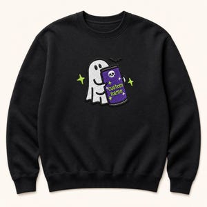 May include: A black crewneck sweatshirt featuring an embroidered design of a white ghost holding a purple beverage can. The can displays a white skull icon, the text "custom name" in green, and small yellow stars. Bright green stars surround the ghost.