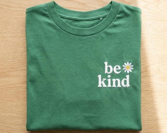 May include: A forest green crew neck t-shirt featuring white embroidered text that reads be kind, with a small white and yellow daisy flower icon positioned to the right of the word be. The shirt has a standard short sleeve design.