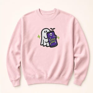 May include: A light pink crewneck sweatshirt featuring an embroidered design of a white ghost holding a purple can with a skull icon and the text custom name. The design includes a small black bat and four lime green four-pointed stars.