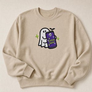 May include: A beige crewneck sweatshirt featuring an embroidered design of a white ghost holding a purple can with a skull icon and the text "custom name" in green, accented by small yellow stars and a black bat silhouette above the can.