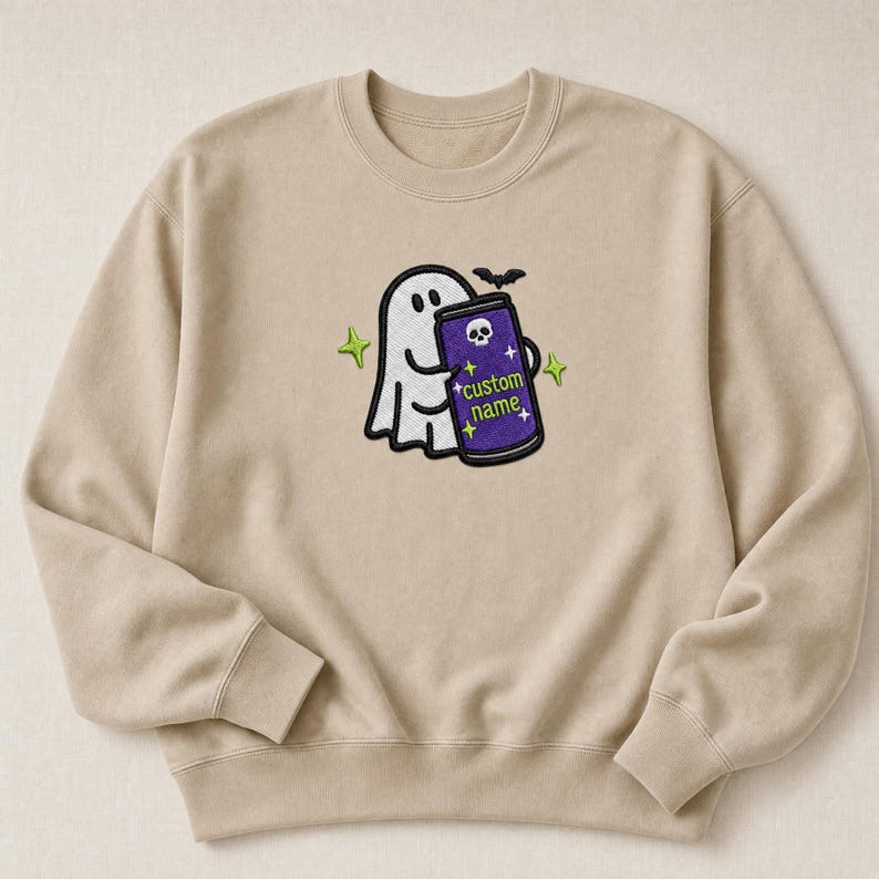 May include: A beige crewneck sweatshirt featuring an embroidered design of a white ghost holding a purple can with a skull icon and the text "custom name" in green, accented by small yellow stars and a black bat silhouette above the can.