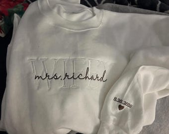 May include: A white long-sleeve sweatshirt featuring the embroidered text WIFEY in large block letters with the cursive text mrs. richard layered over it. The sleeve includes the embroidered date 26.8.2026 above a small brown heart icon.