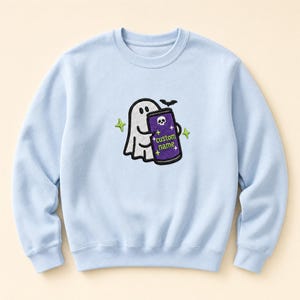 May include: A light blue crewneck sweatshirt featuring a central embroidered graphic of a white ghost holding a purple beverage can. The can displays a small white skull icon, the text custom name, and several small yellow four-pointed stars.