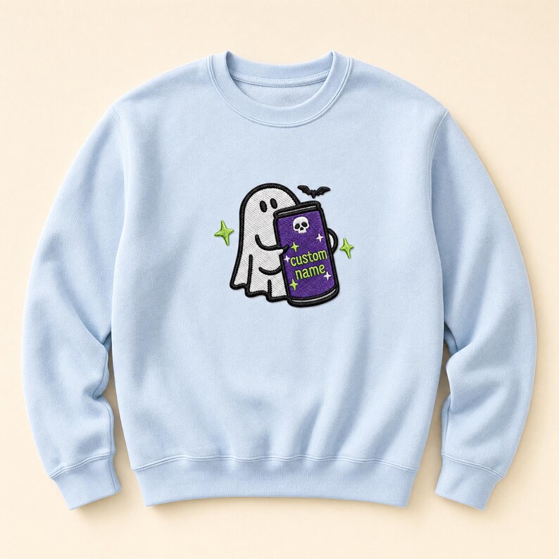 May include: A light blue crewneck sweatshirt featuring a central embroidered graphic of a white ghost holding a purple beverage can. The can displays a small white skull icon, the text custom name, and several small yellow four-pointed stars.