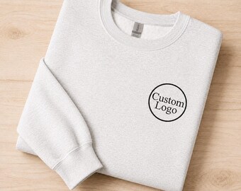 May include: A light grey crewneck sweatshirt featuring a circular black outline on the left chest containing the black text Custom Logo. The garment has long sleeves with ribbed cuffs and a ribbed neckline, showcasing a clean and simple design.