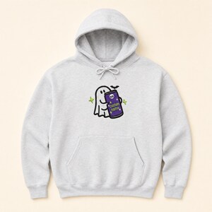 May include: A light grey hooded sweatshirt featuring a central embroidered graphic of a white ghost holding a purple beverage can. The can displays a small skull icon, the text "custom name", and is surrounded by small green star accents.
