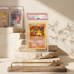 May include: A 1999 Pokemon Game Charizard trading card encased in a clear protective PSA slab. The card features a holographic illustration of the orange dragon-like creature, 120 HP, and the text Charizard, Energy Burn, and Fire Spin.