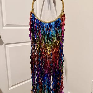 May include: A circular hoop featuring a textured magenta top section and a gold-toned frame. Numerous long, wavy, metallic ribbons in a spectrum of rainbow colors, including blue, red, green, purple, and gold, hang vertically from the bottom half.