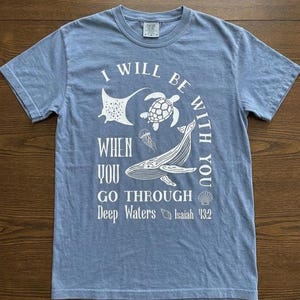 May include: A light blue short-sleeve t-shirt featuring a white graphic print of a whale, sea turtle, stingray, jellyfish, and seashells. The text reads: I WILL BE WITH YOU WHEN YOU GO THROUGH Deep Waters Isaiah 43:2. The shirt has a crew neckline.