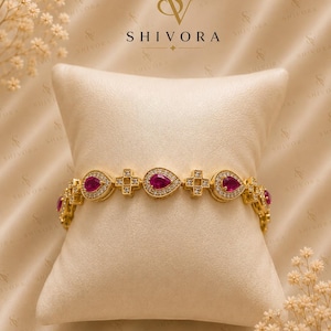 Puede incluir: Una pulsera de tono dorado con piedras de color rosa intenso en forma de pera que alternan con eslabones en forma de cruz, todo incrustado con pequeñas piedras blancas transparentes. La joya se exhibe sobre un cojín crema con la marca SHIVORA.