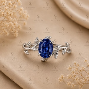 Peut inclure: Une bague argentée ornée d'une pierre précieuse bleu profond de taille ovale au centre. L'anneau présente un motif de feuilles et de vignes métalliques qui s'étend des deux côtés de la pierre, la fixant avec quatre griffes.