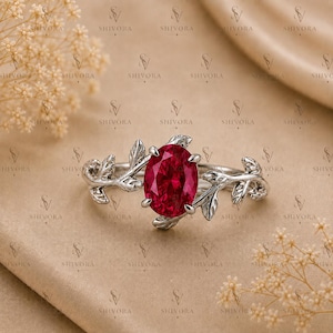 Pode incluir: Um anel de tom prateado com uma pedra preciosa de cor vermelho-profundo de corte oval no centro. A banda apresenta um design de folhas metálicas detalhadas que se estendem de ambos os lados da pedra, criando uma aparência de ramo.
