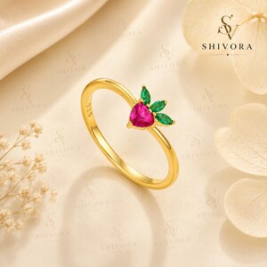 Puede incluir: Un anillo dorado con una piedra preciosa rosa en forma de corazón y tres piedras verdes de talla marquesa dispuestas como hojas. La banda tiene el sello 925, que indica plata de ley. El diseño tiene la apariencia de una fresa con engaste.