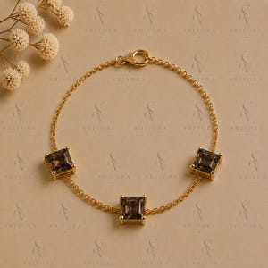 Puede incluir: Una pulsera de cadena dorada con tres piedras preciosas de color marrón oscuro de corte cuadrado en engastes de cuatro garras. La pulsera incluye un cierre de mosquetón y está decorada con un pequeño grupo de flores blancas secas arriba.