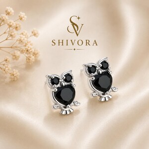 May include: A pair of silver stud earrings shaped like owls, featuring two small round black stones for eyes and a larger heart-shaped black stone for the body. The text SHIVORA is printed above the earrings with a stylized SV logo above it.