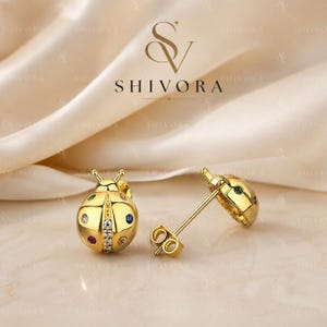 May include: A pair of gold-toned ladybird stud earrings featuring a central row of clear stones and scattered colourful accent stones on the wings. The earrings include post backs and are branded with the text SHIVORA above the jewellery pieces.