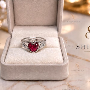 Puede incluir: Un anillo de tono plateado con una piedra preciosa roja en forma de corazón sostenida por dos manos metálicas, presentado dentro de un joyero forrado de terciopelo beige. El nombre de la marca SHIVORA aparece a la derecha de la caja.