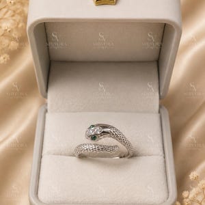 May include: A silver-toned snake-shaped ring featuring a textured scale pattern, two clear stones on the head, and two green stones for eyes, presented inside a light gray rectangular jewelry box with a gold-colored crown emblem on the lid.
