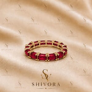 May include: A gold eternity band featuring a continuous row of rectangular, baguette-cut red rubies set in a polished gold metal frame. The ring displays a uniform design with each gemstone secured by small gold prongs, creating a circular shape.