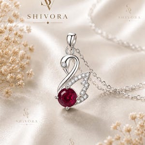 May include: A silver pendant necklace featuring a swan-shaped design with a round, deep red gemstone at the base and small clear stones along the wing. The pendant hangs from a delicate silver chain. The text SHIVORA is visible in the image.