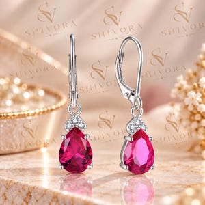 May include: A pair of silver leverback earrings featuring a pear-shaped, deep pink gemstone suspended from a small triangular setting adorned with three tiny, clear, round-cut stones. The metal has a polished finish and a bright, reflective surface.