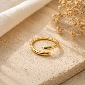 May include: A gold-toned open ring with a smooth, polished finish. The band features a simple, minimalist design with two rounded, tapered ends that do not meet, creating a continuous circular shape with a small gap between the two ends.