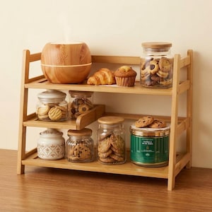 May include: A three-tiered bamboo shelf holding a wood-grain essential oil diffuser, a glass jar of cookies, a croissant, a muffin, and various glass and metal canisters filled with cookies and treats, including a green Vanilla Bean Noel tin.