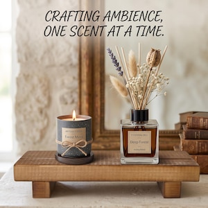 May include: A lit candle in a speckled gray ceramic jar with a twine bow and a glass reed diffuser with dried flowers, both labeled Artisan Scents Forest Moss and Essential Oils Deep Forest, on a wooden riser with the text Crafting ambience, one scent at a time.