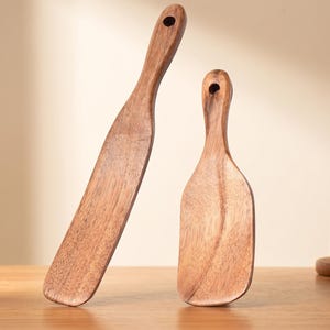 May include: Two wooden kitchen spatulas with visible grain patterns. The taller spatula has a long, narrow blade, while the shorter one features a wider, rounded blade. Both utensils include a circular hole at the top of the handle for hanging.