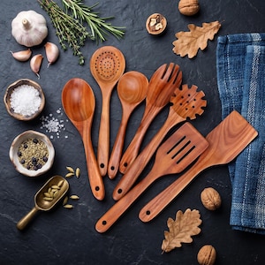 May include: A set of seven wooden kitchen utensils including a slotted spoon, a solid spoon, a pasta fork, and two spatulas, arranged alongside garlic, herbs, spices, walnuts, and a blue cloth napkin on a dark textured surface.